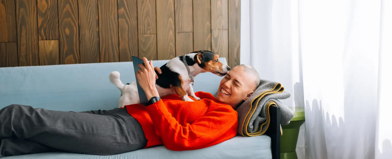 Person in red sweater lying on couch laughing with small dog